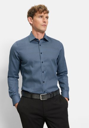 Homme aux cheveux bouclés portant une chemise ajustée bleu foncé à motifs avec des boutons et un pantalon noir avec une ceinture noire, les mains dans les poches.
