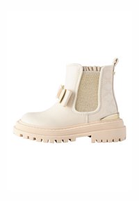 Bottine beige avec un panneau latéral élastique texturé, semelle en caoutchouc, bout arrondi et détail de nœud décoratif à l'avant.