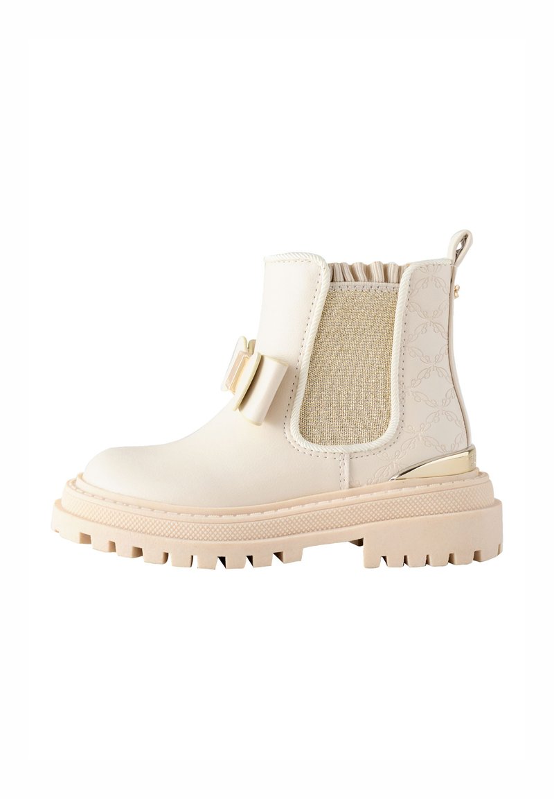 Bottine beige avec un panneau latéral élastique texturé, semelle en caoutchouc, bout arrondi et détail de nœud décoratif à l'avant.