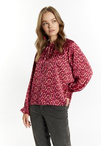 Blusa a maniche lunghe in rosa e rosso con motivo astratto, vestibilità comoda, polsini con volant e scollatura con cordoncino. Abbinata a pantaloni grigio scuro.