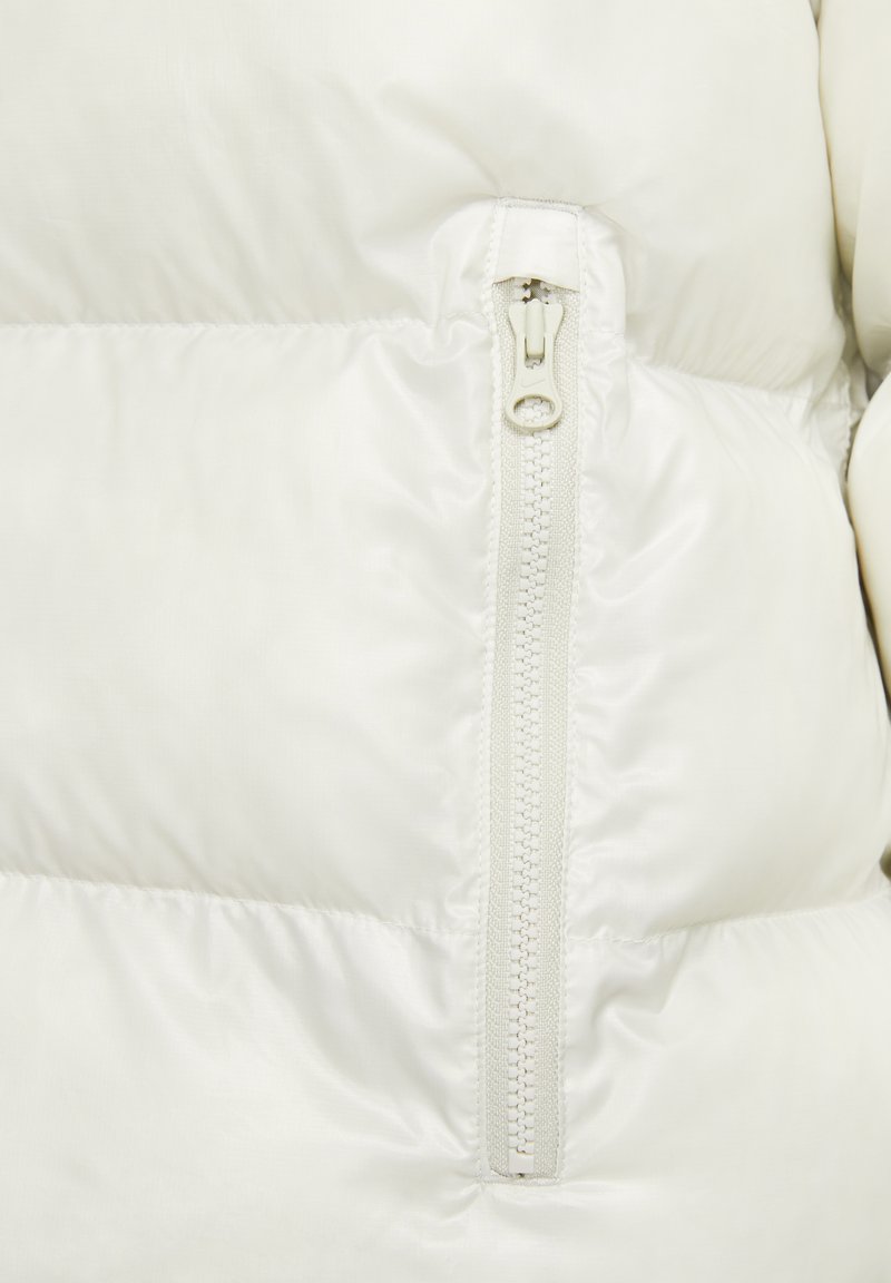 Primer plano de una chaqueta de plumas blanca que muestra una textura lisa y brillante. Presenta un bolsillo con cremallera y una cremallera a juego, destacando un diseño minimalista.