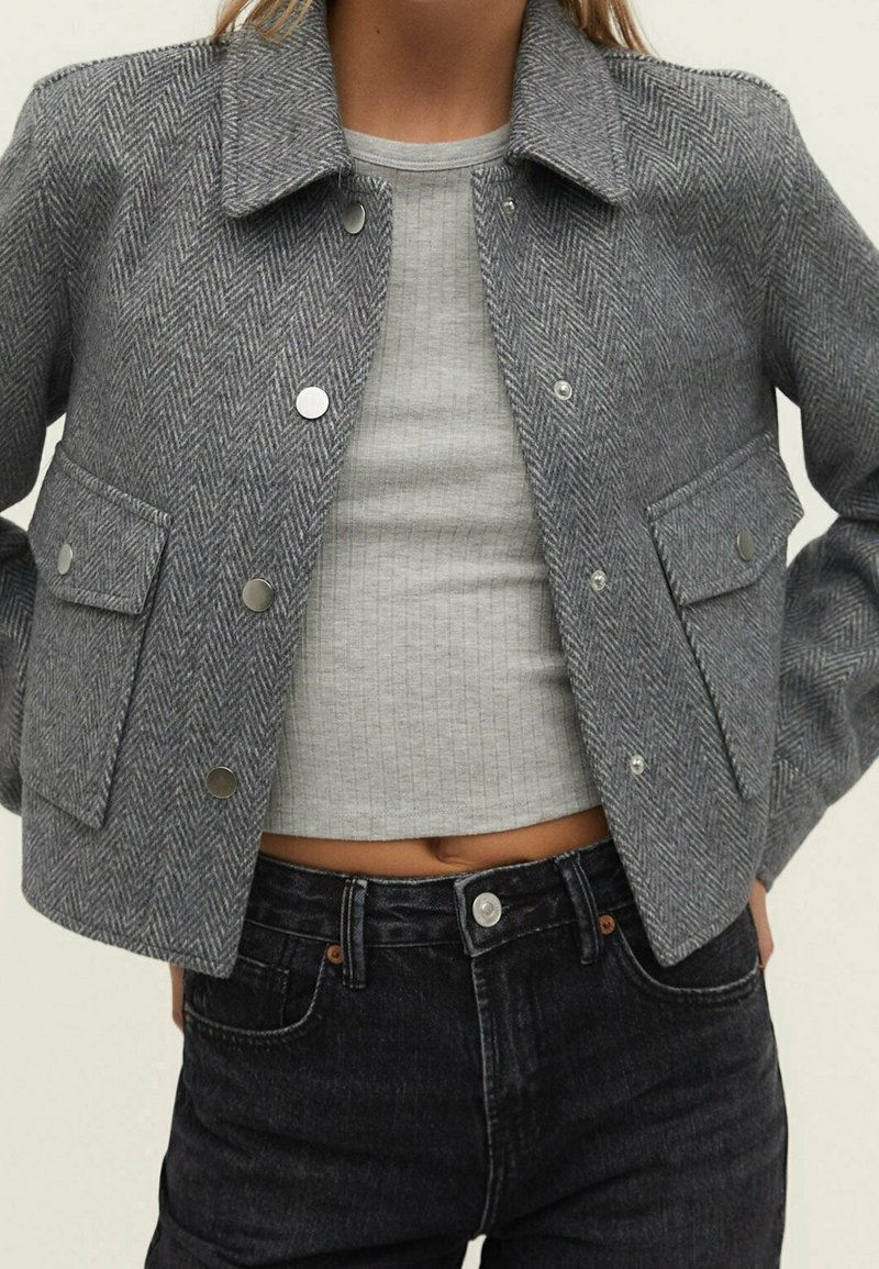 Szara krótka kurtka w wzór jodełki z kołnierzem, srebrnymi napami i dwiema przednimi kieszeniami, noszona na szarym prążkowanym topie i ciemnych dżinsach.