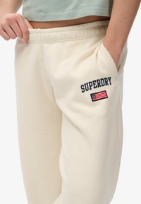 Crème-kleurige sweatpants van zacht materiaal, met een geborduurd "SUPERDRY" logo en vlagdetail op de linker zak.