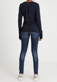 Person som står med ryggen mot kameran, iklädd en marinblå långärmad skjorta med små prickar, åtsittande blå jeans och vita sneakers med rosa detaljer.