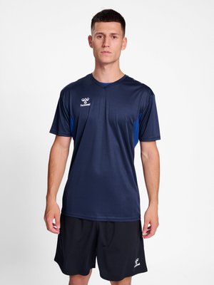 Młody mężczyzna stojący, ubrany w granatową sportową koszulkę i czarne spodenki z logo hummel, zwrócony przodem na białym tle.