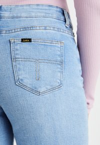 Lyseblå denimjeans har en diamantskåret baklomme med gul søm og en svart "Lois"-label. Glatt tekstur, høyt liv-design.
