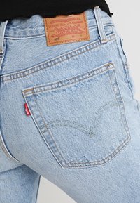 Světlé modré džíny z denimu s nášivkou označenou "501", dvěma zadními kapsami a červeným logem na levé kapse. Opotřebené detaily poblíž švů.