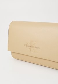 Portafoglio sintetico beige e liscio con design pieghevole, caratterizzato da logo "CK" in rilievo e testo "Calvin Klein Jeans". Cuciture a vista, minimalista.