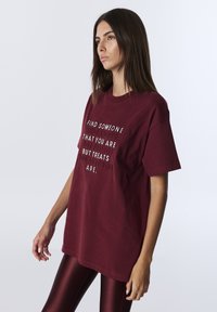 Bordowa bawełniana koszulka z krótkim rękawem i luźnym krojem. Z przodu znajduje się wyrazisty nadruk w kontrastowych kolorach. Noszona z błyszczącymi, dopasowanymi legginsami.