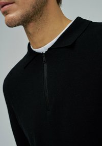 Zbliżenie na mężczyznę w czarnej, teksturowanej bluzie na zamek z kołnierzykiem, która odsłania białą koszulę pod spodem.