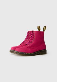 Bottines en suède rose vif avec des lacets assortis, des tirettes jaunes et noires, et une semelle contrastante noire avec des surpiqures jaunes.