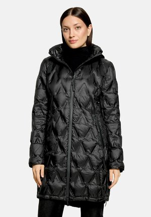 Femme portant une longue veste matelassée noire avec une fermeture éclair à l'avant, un col montant et des poches latérales zippées, posant devant un fond clair uni.