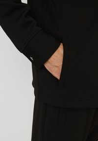 Person's hand partially inside a black jacket pocket, wearing matching black pants, against a plain light background.