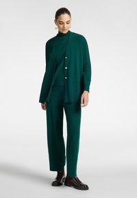 Nainen, joka on pukeutunut tummanvihreisiin leveälahkeisiin housuihin, yhteensopivaan poolopaitaan, pitkään napilla varustettuun neuletakkiin ja mustiin paksupohjaisiin saappaisiin, seisoo valkoista taustaa vasten.