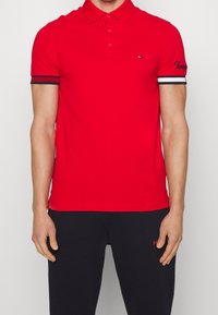 Rød polo shirt lavet af glat stof, med en klassisk krave, tre knapper og sorte og hvide stribede detaljer på ærmerne. Broderet logo.