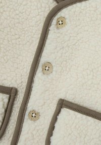 Cremfarbener Fleece-Stoff mit strukturierter Oberfläche, in Braun eingefaßt. Verfügt über drei runde Knöpfe und zwei vordere Taschen.