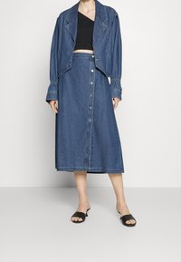 Jeansjacka med en figursydd design, bred krage och kort längd, ihop med en lång jeanskjol med knapprad. Svarta slip-on sandaler.