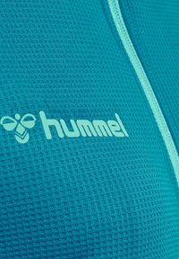 Teal sportstof met een gestructureerd patroon; heeft een lichtblauwe rits en een lichtblauw "hummel" logo in het midden.