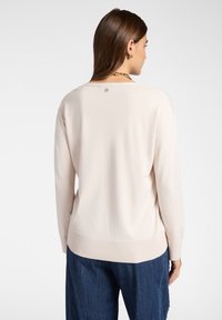 Una donna con capelli castani indossa un maglione crema a maniche lunghe e pantaloni blu, girata di spalle, mostrando il retro del maglione e un piccolo logo vicino al collo.