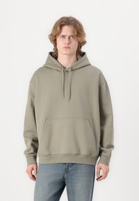 Olive green, oversized hoodie in soft fabric with a kangaroo pocket, drawstring hood, and ribbed cuffs and hem. Worn with light blue jeans.