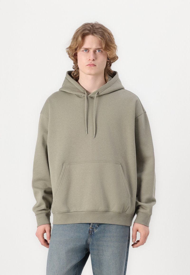 Olive green, oversized hoodie in soft fabric with a kangaroo pocket, drawstring hood, and ribbed cuffs and hem. Worn with light blue jeans.