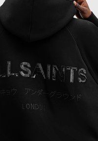 Black hoodie featuring "ALLSAINTS" in shiny sequins, with embroidered text in different languages below. Soft fabric, relaxed fit, and a hood.