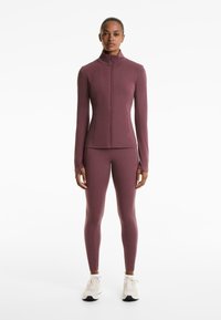 Haut de sport à manches longues et fermeture éclair, avec un legging assorti en tissu mauve foncé, au design ajusté, texture lisse, coutures minimales. Baskets blanches.