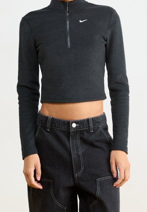 Person iført en sort cropped Nike fleece med halv lynlås og sort højttaljet bukser med synlige hvide sømme og lommer.