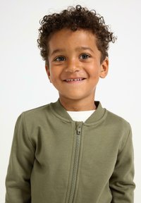 Olivengrüne Kapuzenjacke mit Rundhalsausschnitt und weicher Textur, über einem weißen Hemd getragen. Das Modell hat lockige Haare und einen neutralen Gesichtsausdruck.