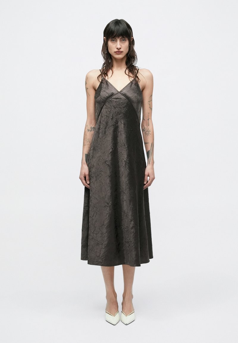 Tumši brūnas krāsas bezroku kleita ar saburzītu tekstūru, V veida kakla izgriezumu un A līnijas siluetu. Valkāta ar gaiši krāsainām smailām augstpapēžu kurpēm.