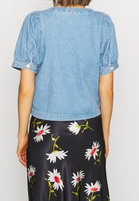 Veste courte en denim bleu clair avec manches bouffantes et ornements de boutons, portée sur une jupe noire à motif floral.