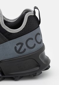 Baskets noires et grises avec une surface texturée, un col rembourré et un logo bien visible. Elles présentent une semelle robuste avec un dessin antidérapant.