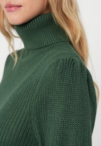 Suéter verde de gola alta tricotado com textura ribana, costuras estruturadas nos ombros e um silhueta ajustada.