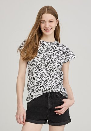 Blusa bianca con motivo floreale nero, maniche corte e collo rotondo. Abbinata a pantaloni corti di jeans neri a vita alta.
