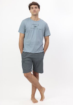 Joven de pie descalzo con camiseta azul claro "Check-in Antonio Miro" y pantalones cortos oscuros con rayas, manos en los bolsillos, sobre fondo blanco.