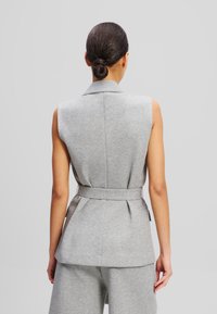 Grijze mouwloze blazer met gestructureerde schouders, brede taille en geplooide achterkant. Gemaakt van een zachte, gestructureerde stof voor een casual uitstraling.