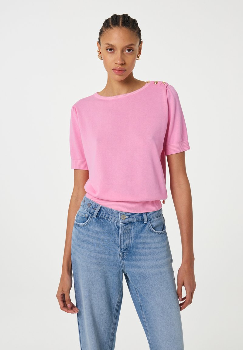 Roze gebreide trui met korte mouwen, ronde halslijn en decoratieve gouden knopen op de schouder, gecombineerd met lichtblauwe denim jeans.