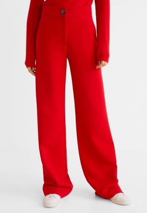 Femme portant un pantalon large rouge vif et un haut à manches longues assorti, associée à des baskets blanches, debout devant un fond uni.