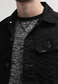 Giacca in denim nero con bottoni a pressione e tasche a toppa, indossata sopra un maglione in maglia grigia testurizzata con collo rotondo.