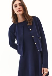 Granatowy kardigan z guzikami w złotym odcieniu, prążkowanymi mankietami i dwiema przednimi kieszeniami, w zestawie z dopasowaną sukienką w podobnym kolorze i materiale.