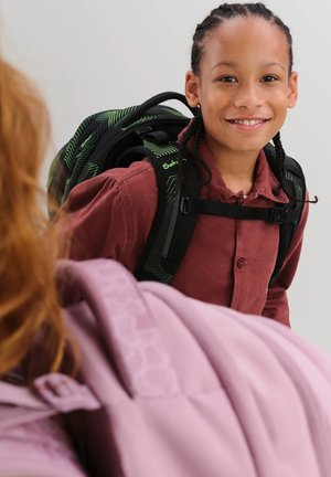 Lächendes Kind mit geflochtenen Haaren, das ein dunkelrotes Hemd und einen grünen Rucksack trägt und einer anderen Person mit einem hellrosa Rucksack gegenübersteht.