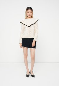 Woman wearing cream cable-knit sweater with large ruffled collar and black trim, paired with black mini skirt and black pointed flats.