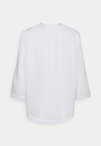 Blouse blanche à manches longues avec de doux plis au niveau de l'encolure, présentée de dos sur un fond uni.