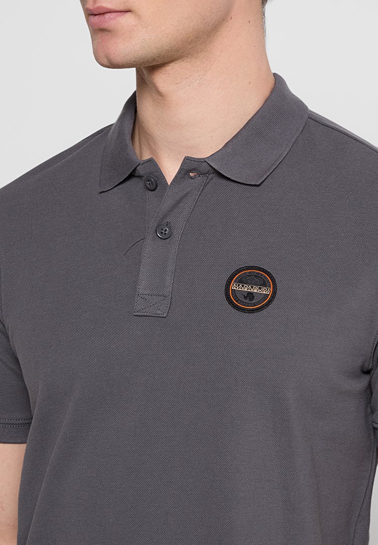 Graues Poloshirt aus strukturiertem Stoff, mit einer Knopfleiste mit zwei Knöpfen, umgelegtem Kragen und einem runden Logo-Patch mit orangefarbenen Akzenten.