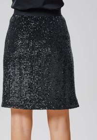 Jupe noire en sequins avec une surface texturée, longueur courte et taille élastique. Des sequins brillants créent un motif réfléchissant sur l'ensemble.