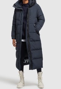 Lång, mörkblå puffermantel med huva, med ett kviltat mönster, två sidofickor och dragkedjestängning. Bärs över svarta leggins och ljusa stövlar.