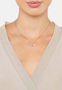 Sølv halskæde med en libelle vedhæng udsmykket med klare ædelsten, hvilende på en delikat kæde. Båret over et lyst stof.