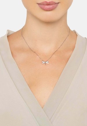 Silver necklace featuring a dragonfly pendant adorned with clear gemstones, resting on a delicate chain. Worn against a light fabric.