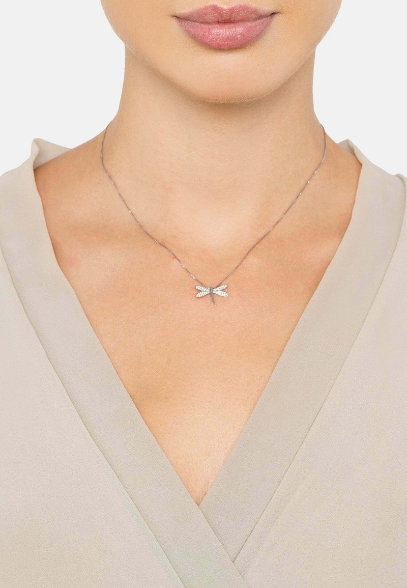 Sølv halskæde med en libelle vedhæng udsmykket med klare ædelsten, hvilende på en delikat kæde. Båret over et lyst stof.