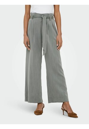 Ženska, ki nosi sive široke hlače z visokim pasom in tkanim pasom, zavezanim v pasu, ter rjave čevlje z odprtim zadnjim delom in peto.